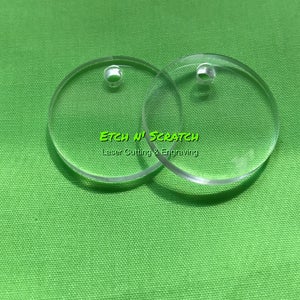 May include: Two clear, circular acrylic blanks with small holes for crafting or jewelry making. The text "Etch N' Scratch Laser Cutting & Engraving" is visible in green. The blanks are on a green background.