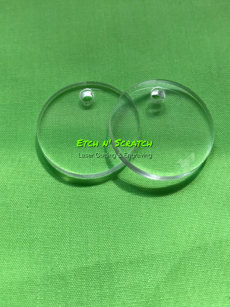 May include: Two clear, circular acrylic blanks with small holes for crafting or jewelry making. The text "Etch N' Scratch Laser Cutting & Engraving" is visible in green. The blanks are on a green background.