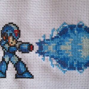May include: Cross-stitch artwork featuring a blue robot character firing a blue energy blast. The robot is depicted in a pixelated style, reminiscent of classic video games.