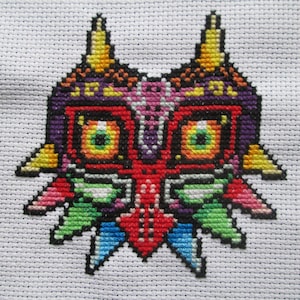 May include: A cross-stitch of a colorful mask with a detailed design. The mask features a red, purple, yellow, green, and blue color scheme. The mask is set against a white fabric background.