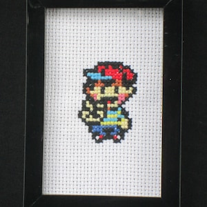 May include: A framed cross-stitch of a pixelated character wearing a red hat, blue shirt, and yellow pants. The character is standing with their arms raised.