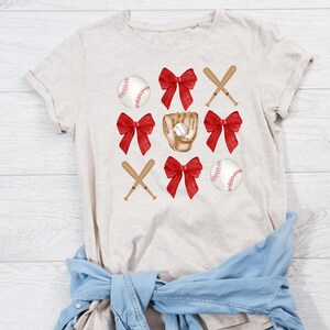 May include: A beige t-shirt with a baseball theme. The design features red bows, baseball bats, baseballs, and a baseball glove.
