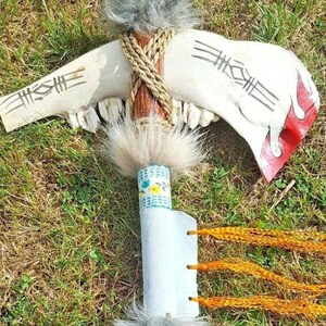 May include: A handmade Native American style tomahawk with a white bone head, brown wooden handle, and grey fur accents. The handle is wrapped with brown twine and decorated with beads and leather. The tomahawk is lying on a bed of green grass.