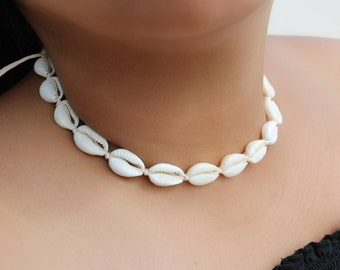 May include: A white cowrie shell choker necklace. The shells are strung together with a cream-coloured cord, creating a delicate and natural look. The necklace is worn around the neck, showcasing the shells' smooth texture and organic shape.