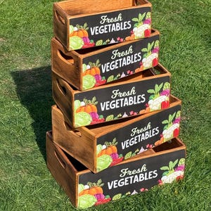 Può includere: Una pila di cinque casse di legno con la scritta "Fresh Vegetables" sul davanti. Ogni cassa ha un fronte nero con illustrazioni colorate di verdure. Le casse sono in legno marrone.