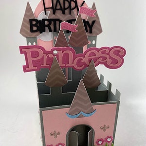 May include: A pink and gray paper castle with the words "Happy Birthday Princess" on top. The castle has brown chevron-patterned turrets and pink flags. The base of the castle is pink with a gray door and decorative flowers.