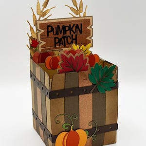 May include: A decorative wooden pumpkin patch box with a sign, filled with pumpkins and fall leaves. The box is brown with vertical stripes and metal bands. A large orange pumpkin is on the front.