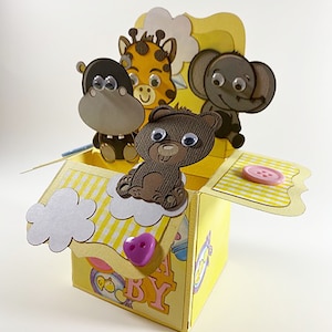 May include: A yellow pop-up box with a baby theme, featuring a hippo, giraffe, bear, and elephant. The box has a striped roof, cloud accents, and a purple heart-shaped button closure. The text "A BABY" is visible on the side.