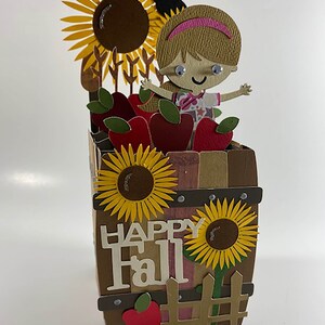 May include: A papercraft fall-themed diorama featuring a girl, sunflowers, and apples in a barrel. The barrel has the words "Happy Fall" on the front. A black crow sits atop the sunflowers. The diorama is made of various shades of brown, yellow, red, and green.