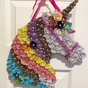 May include: A colorful unicorn head wall hanging made of paper flowers in shades of blue, pink, yellow, purple, and brown. The unicorn has a pearl-studded mane and a horn made of rolled paper. A pink ribbon is attached for hanging.
