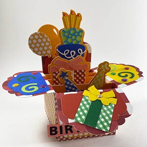May include: A colorful pop-up birthday card featuring a layered cake, wrapped gifts, balloons, and party hats. The card has a red and white checkered pattern and the word "BIR" is visible. The cake has blue and green layers.