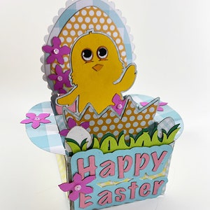 May include: A handmade Easter-themed papercraft featuring a yellow chick hatching from a decorated egg. The design includes pink flowers, green grass, and the words "Happy Easter" in pink letters on a blue background. The overall design is a 3D pop-up card.