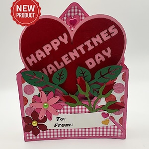 May include: A Valentine's Day card with a red heart that says "HAPPY VALENTINES DAY". The card is pink with a white and pink gingham pattern and features decorative flowers and leaves. A "NEW PRODUCT" sticker is in the upper left corner. There is a space to write "To:" and "From:"