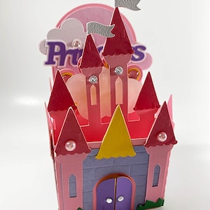 May include: A pink and purple paper castle with the word "Princess" on top. The castle has red turrets, a yellow roof, and a purple door. Silver flags and decorative gems add detail to the design. The castle is a decorative item.