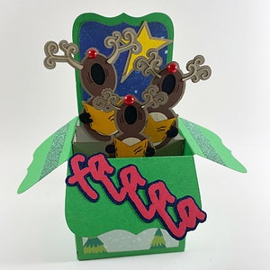 May include: A pop-up Christmas card featuring three reindeer singing carols. The reindeer are brown with red noses and hold yellow songbooks. The card is green with the words "fa la la" in red and blue.
