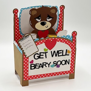 May include: A handmade get well card featuring a brown bear in a bed. The bed frame is red with white polka dots. The card reads "Get Well Beary Soon" with heart accents. A clipboard and a heart are also included.
