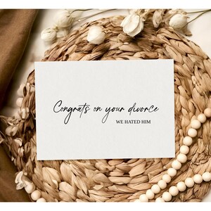 May include: A white card with black text that reads "Congrats on your divorce. We hated him." The card is sitting on a woven straw mat with white beads.