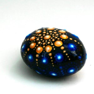 May include: A smooth, oval-shaped stone painted with a black base color. The surface features a mandala-style dot art pattern with a central cluster of gold dots radiating outward, surrounded by scattered, vibrant blue dots of varying sizes.