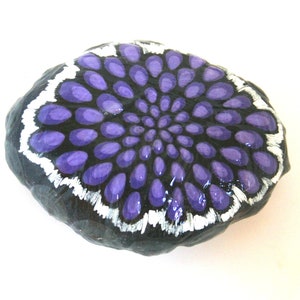 Pode incluir: Uma pedra pintada com um desenho de mandala roxo e branco. A pedra &eacute; preta e tem uma forma redonda e lisa.