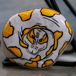 May include: A hand-painted, rounded stone featuring a snake design. The stone is white with yellow and black accents, and the snake's face is visible in the center. The artwork is displayed on a gray surface.