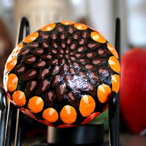 May include: A painted rock with a black, brown, orange, and white geometric design. The rock is on a black metal stand.