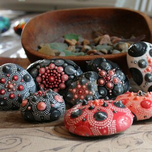 May include: A collection of eight hand-painted stones with intricate designs. The stones are painted in shades of black, white, and pink with a variety of patterns.