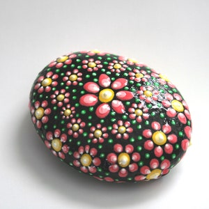 Puede incluir: Una piedra pintada con fondo negro y flores rosas y amarillas. La piedra es lisa y de forma ovalada.