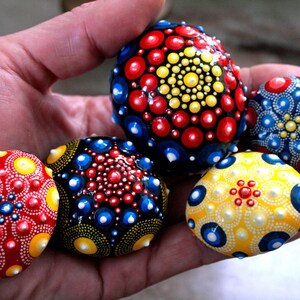 Puede incluir: Cinco piedras pintadas con intrincados patrones de puntos en rojo, amarillo, azul y blanco. Las piedras están dispuestas en una mano, con la piedra más grande en el centro.