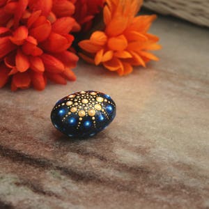 May include: A smooth, oval-shaped stone painted with a dark blue base and a mandala-style pattern of small, raised dots in shades of gold, light blue, and white, arranged in a symmetrical, radiating design that covers the top surface.