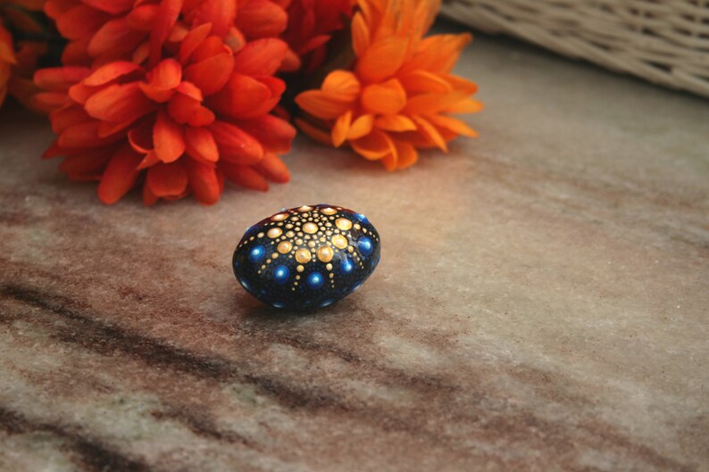 May include: A smooth, oval-shaped stone painted with a dark blue base and a mandala-style pattern of small, raised dots in shades of gold, light blue, and white, arranged in a symmetrical, radiating design that covers the top surface.