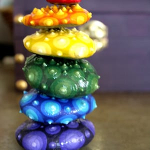 May include: A stack of six colorful, hand-painted, pebble-shaped stones. The stones are arranged in a vertical tower, each a different color: red, orange, yellow, green, blue, and purple. Each stone has a textured, bumpy surface.