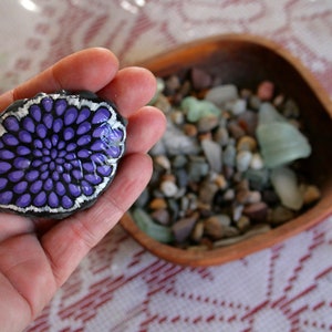 Pode incluir: Uma m&atilde;o segura uma pedra pintada com um desenho de flor roxa e branca. A pedra est&aacute; sobre uma mesa com uma tigela de pequenas pedras.