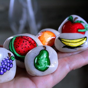 Puede incluir: Seis piedras pintadas con diferentes dise&ntilde;os de frutas. Las piedras est&aacute;n pintadas con una sand&iacute;a, una pera, un racimo de uvas, un pl&aacute;tano, una naranja y una manzana con un gusano.