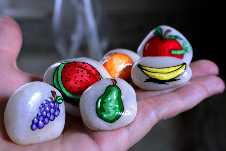 Puede incluir: Seis piedras pintadas con diferentes dise&ntilde;os de frutas. Las piedras est&aacute;n pintadas con una sand&iacute;a, una pera, un racimo de uvas, un pl&aacute;tano, una naranja y una manzana con un gusano.