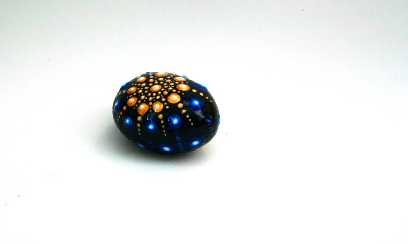 May include: A smooth, oval-shaped stone painted with a black base color. The surface features a mandala-style dot art pattern with a central cluster of gold dots radiating outward, surrounded by scattered, vibrant blue dots of varying sizes.