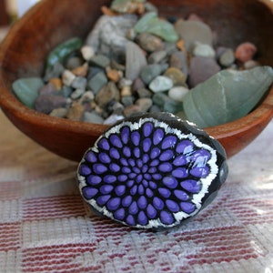 Pode incluir: Uma pedra pintada com um desenho de mandala roxo e branco. A pedra est&aacute; sobre uma toalha de mesa quadriculada branca e vermelha.
