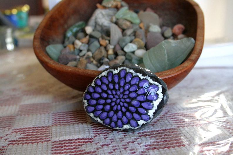 Pode incluir: Uma pedra pintada com um desenho de mandala roxo e branco. A pedra est&aacute; sobre uma toalha de mesa quadriculada branca e vermelha.