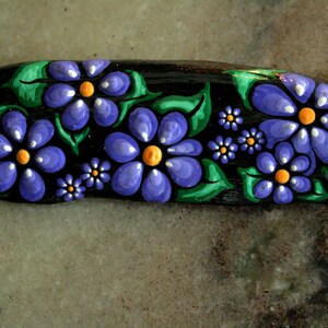 Puede incluir: Una piedra negra oblonga pintada a mano con un diseño floral. Las flores son moradas con centros amarillos y detalles blancos, rodeadas de hojas verdes. La obra de arte es detallada y vibrante.