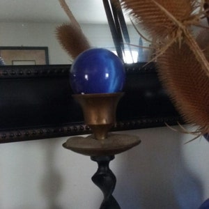 May include: A dark blue glass sphere rests atop a brass candle holder. The holder features a wide, flat circular base plate and a slender, twisted black metal stem that extends downward, supporting the polished brass cup holding the blue orb.
