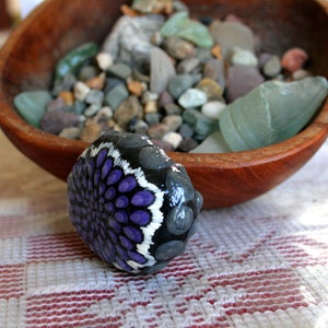 Pode incluir: Uma pedra pintada &agrave; m&atilde;o com um desenho de mandala roxo e branco. A pedra est&aacute; sobre uma toalha de mesa quadriculada branca e vermelha.