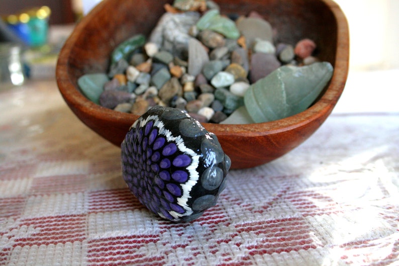 Pode incluir: Uma pedra pintada &agrave; m&atilde;o com um desenho de mandala roxo e branco. A pedra est&aacute; sobre uma toalha de mesa quadriculada branca e vermelha.