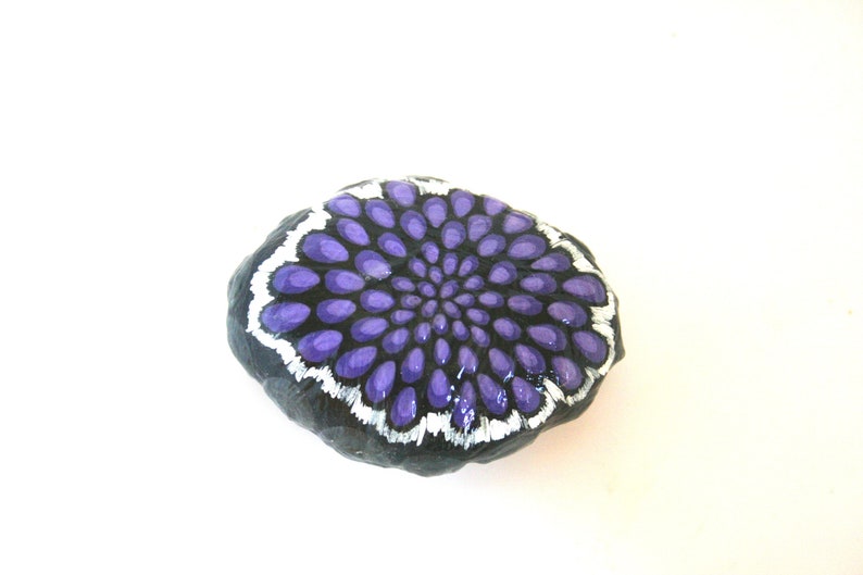 Pode incluir: Uma pedra pintada com um desenho de mandala roxo e branco. A pedra &eacute; preta e tem uma forma redonda e lisa.