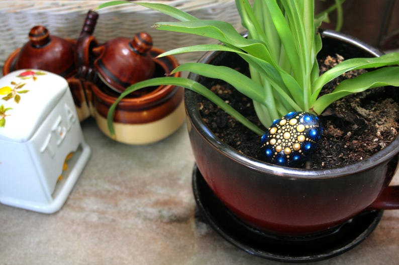 May include: A dark red and black ceramic planter containing a green plant and a small, smooth, oval-shaped stone decorated with a blue and gold mandala dot pattern. Nearby sits a white ceramic tea container and a brown ceramic condiment set.