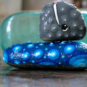 May include: A stack of three painted rocks. The top rock is gray and has a white line and black eyes. The middle rock is blue with white dots. The bottom rock is blue with white circles.