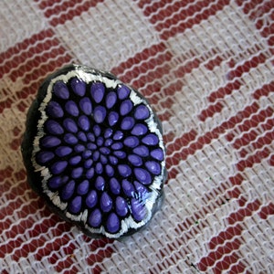 Pode incluir: Uma pedra pintada com um desenho de flor roxa e branca. A pedra &eacute; redonda e tem uma superf&iacute;cie lisa.