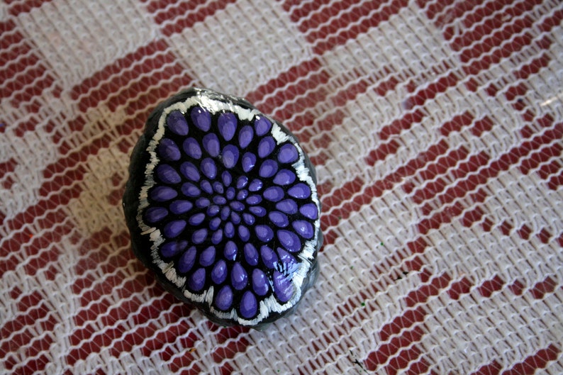 Pode incluir: Uma pedra pintada com um desenho de flor roxa e branca. A pedra &eacute; redonda e tem uma superf&iacute;cie lisa.