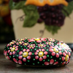 Puede incluir: Una piedra pintada de negro con flores rosas y verdes, centros blancos y puntos amarillos. La piedra est&aacute; pintada en un estilo mandala de puntos.