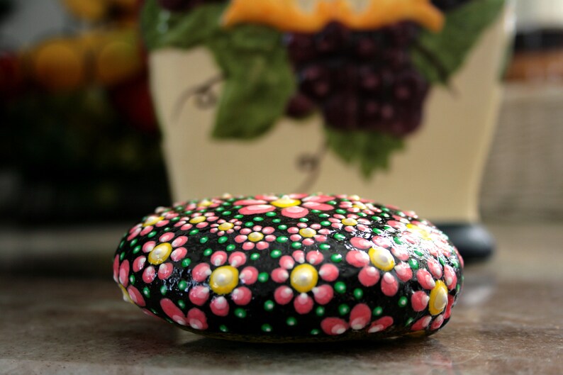 Puede incluir: Una piedra pintada de negro con flores rosas y verdes, centros blancos y puntos amarillos. La piedra est&aacute; pintada en un estilo mandala de puntos.