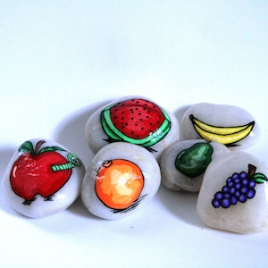 Puede incluir: Seis piedras pintadas, cada una representando una fruta diferente: una manzana roja con un gusano verde, una pera verde, un pl&aacute;tano amarillo, una sand&iacute;a roja, un racimo de uvas moradas y una naranja naranja.