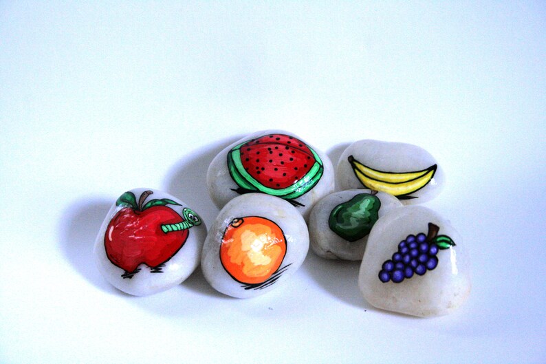 Puede incluir: Seis piedras pintadas, cada una representando una fruta diferente: una manzana roja con un gusano verde, una pera verde, un pl&aacute;tano amarillo, una sand&iacute;a roja, un racimo de uvas moradas y una naranja naranja.